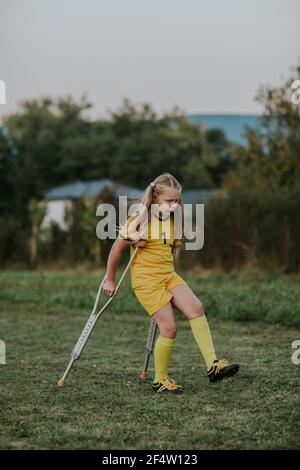 Mädchen mit Krücken zu Fuß entlang Fußballplatz. Volle Länge der verletzten Mädchen Torhüter in gelben Fußballkleid auf Krücken. Stockfoto