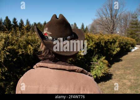 Eine Frau mit kurzen braunen Haaren trägt einen braunen Cattleman Cowboy Hut mit einer Feder und dekorativen Gürtel befestigt. Blick auf Bäume. Stockfoto