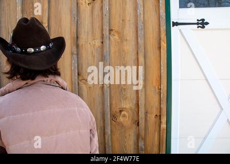 Eine Frau mit kurzen braunen Haaren, die einen braunen Cowboyhut mit einer Feder und einem dekorativen Gürtel trägt, der außerhalb einer Holzscheune und einer Garagentür angebracht ist. Stockfoto