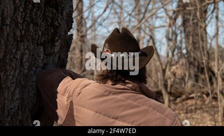 Eine Frau mit kurzen braunen Haaren trägt einen braunen Cattleman Cowboy Hut mit einer Feder und dekorativen Gürtel befestigt. Lehnt sich an einen Baum. Stockfoto