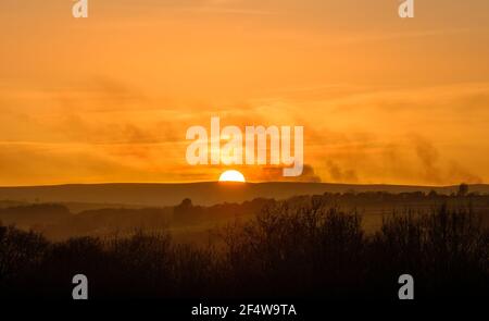 Nebliger orangefarbener Sonnenuntergang über den North Durham Moors am östlichen Rand der Pennines. Rauchen von brennender Heide, die über die untergehende Sonne driftet Stockfoto