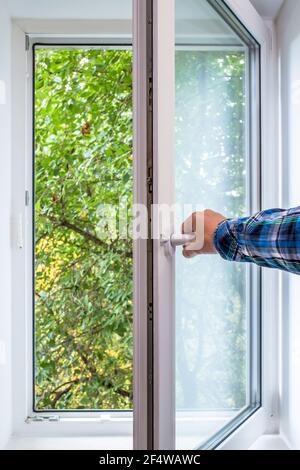 Man Hand öffnet ein PVC-Fenster, von dem aus man einen Baum in einer ökologisch sauberen Gegend der Stadt, mit frischer Luft sehen kann. Stockfoto