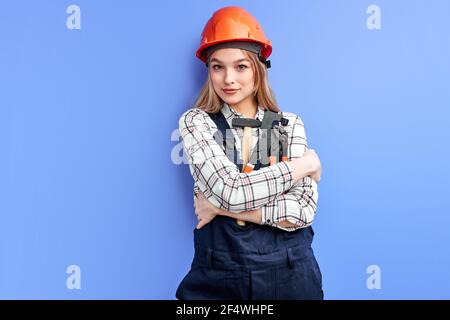 Architektin Frau trägt orange Helm und blauen Overall halten Schlüssel, Hammer und andere Werkzeuge in den Händen isoliert über blauen Studio Hintergrund, Porträt Stockfoto