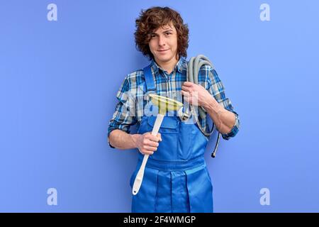 Männlicher Klempner hält Kolben und lächelt an der Kamera auf blauem Hintergrund, Porträt von jungen kaukasischen Kerl mit lockigem Haar in blauen Uniform Schürze gekleidet p Stockfoto