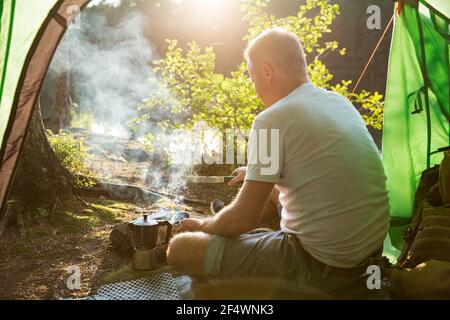 Mann, der Kaffee mit einer Espressomaschine am Lagerfeuer im Wald am Ufer eines Sees macht, im Zelt sitzt, ein Feuer macht, garibert. Glückliches Isolationskonzept. Stockfoto