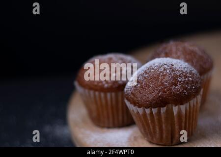 Drei Muffins mit Puderzucker bedeckt auf Holzständer isoliert auf schwarzem dunklen Hintergrund. Bäckerei süß Stockfoto