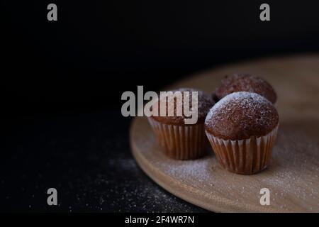 Drei Muffins mit Puderzucker bedeckt auf Holzständer isoliert auf schwarzem dunklen Hintergrund. Bäckerei süß Stockfoto