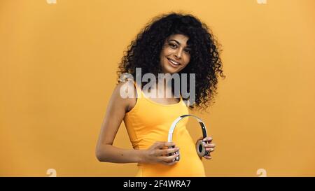 Schwangere hispanische Frau hält Kopfhörer in der Nähe Bauch isoliert auf gelb Stockfoto