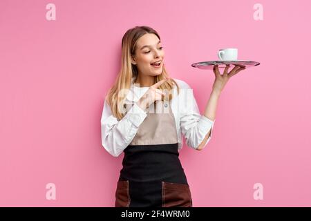 Kellnerin Frau in Schürze hält Tablett mit Tasse Kaffee, Blick mit Überraschung Gesicht zeigt Finger auf Tasse. Isoliert rosa Studio Hintergrund, Porträt Stockfoto