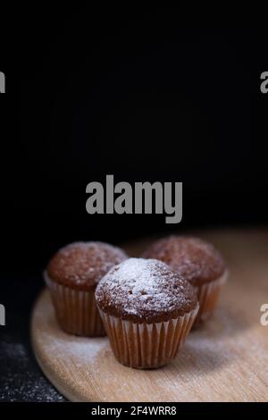 Drei Muffins mit Puderzucker bedeckt auf Holzständer isoliert auf schwarzem dunklen Hintergrund. Bäckerei süß Stockfoto