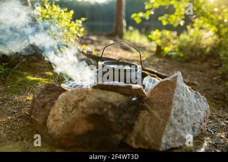 Mann, der im Campingkessel am Lagerfeuer im Wald am Ufer eines Sees Kaffee macht, im Zelt sitzt, ein Feuer macht, am Ufer eines Sees gieren. Glückliches Isolationskonzept. Stockfoto