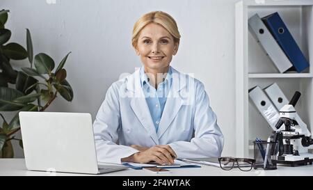 Glücklicher Arzt lächelt an der Kamera, während er in der Nähe des Laptops sitzt Arbeitsplatz Stockfoto