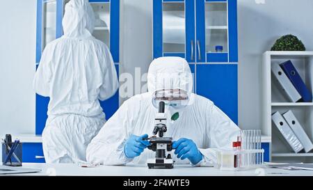 Wissenschaftler in Gefahrgut Anzug arbeiten mit Mikroskop in der Nähe Kollegen in Labor Stockfoto