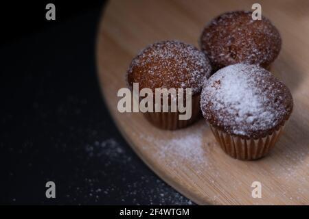 Drei Muffins mit Puderzucker bedeckt auf Holzständer isoliert auf schwarzem dunklen Hintergrund. Bäckerei süß Stockfoto