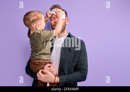 Junger Vater in Augenmaske zum Schlafen, Stand halten Kind Junge in den Händen, wollen am Morgen vor dem Arbeitstag schlafen. Isoliert auf lila Hintergrund Stockfoto