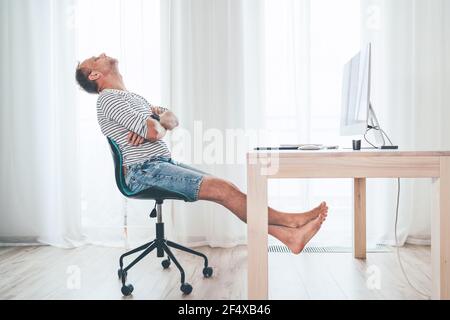 Ein Mann mittleren Alters, der sich auf einem Drehstuhl neben dem Tisch mit Computer entspannt, während er im Wohnzimmer Fernsehen schaut. Er streckt barfuß hinkt und ruht Stockfoto