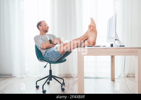 Ein Mann mittleren Alters in einer Brille, der einen modernen Computer in seinem Wohnzimmer beobachtet. Er legte ihre barfuss Lags auf den Tisch und erholte sich. Video-Streaming ein Stockfoto