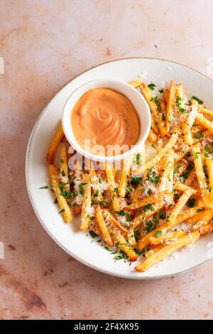 Teller mit goldenen knusprigen Pommes frites mit Parmesankäse und Petersilie und einer kleinen Schüssel rosa Mayonnaise auf der Seite gekrönt. Stockfoto