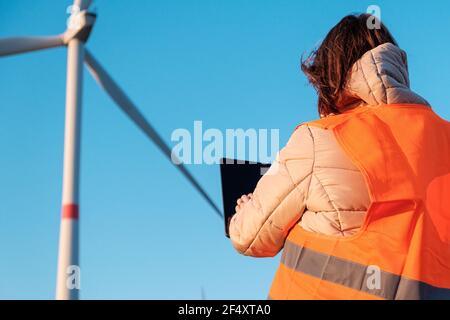 Eine Ingenieurin, die eine Tablette in der Hand hält und im Windturbinenpark auf dem Feld in der orangefarbenen vesta arbeitet Stockfoto