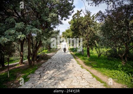 Filopappou Hill, Athen, Griechenland. Historisches Wahrzeichen. Philopappos Waldhintergrund. Gepflasterter Pfad unter dem antiken griechischen Akropolis-Felsen, zwischen Bäumen. Tag, Stockfoto