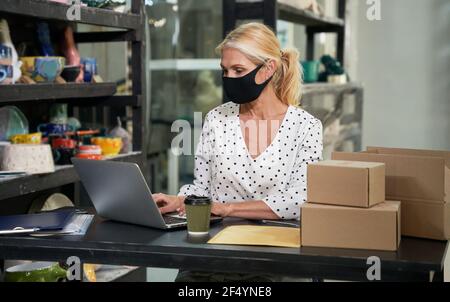 Pandemie. Beschäftigte ältere Frau, Inhaber eines kleinen Unternehmens, die eine Schutzmaske trägt, die am Laptop arbeitet und Bestellungen entgegennimmt, während sie am Schreibtisch sitzt Stockfoto