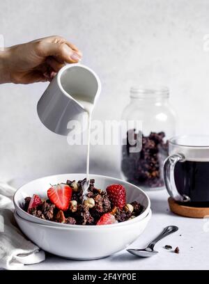 Frühstücksszene: Hausgemachtes Müsli aus Schokolade und Nüssen, serviert in einer Schüssel mit frischen Erdbeeren und Milch auf pflanzlicher Basis, die darüber gegossen wird. Stockfoto