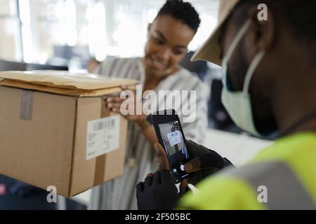 Courier in Gesichtsmaske mit Smartphone-Scan-Paket in Büro Stockfoto
