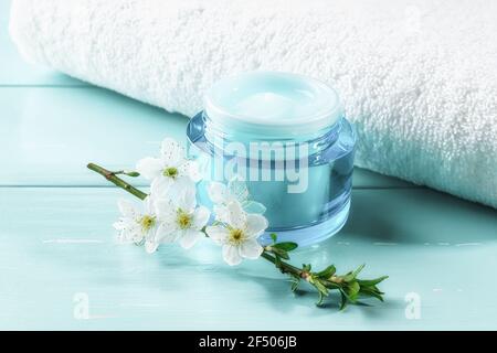 Glas feuchtigkeitsspendende Gesichtsgel und Kirschblüte in der Nähe weißes Handtuch über blaue Holzoberfläche. Blaue Behälter feuchtigkeitsspendende Gesichtscreme. Schönheit, Hautpflege Stockfoto