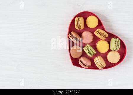 Mehrfarbige französische Makronen in einer herzförmigen Box Draufsicht auf weißem Holzhintergrund. Französische Meringue-Keksmakarons für den Urlaub. Stockfoto