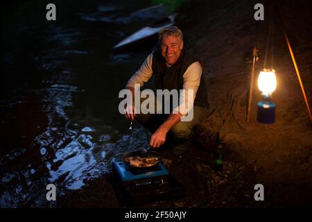 Portrait glücklicher Mann, der auf dem Campingkocher am Flussufer kocht Nacht Stockfoto