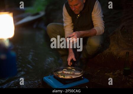 Mann kocht frisch gefangenen Fisch auf Campingkocher in der Nacht Stockfoto