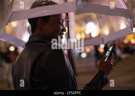 Glückliche junge Frau mit Smartphone unter Regenschirm in der Nacht Stockfoto