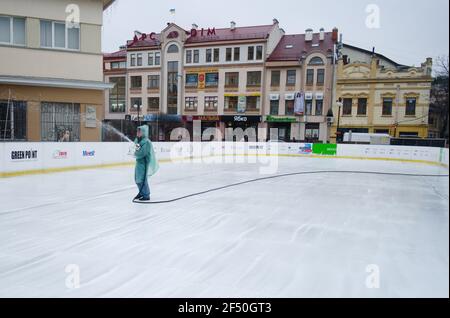 Ivano-Frankivsk, Ukraine - November, 2019: Mann im Regenmantel füllt Eisbahn mit Wasser aus Schlauch. Öffentliche Eisbahn in der Nähe. Stockfoto