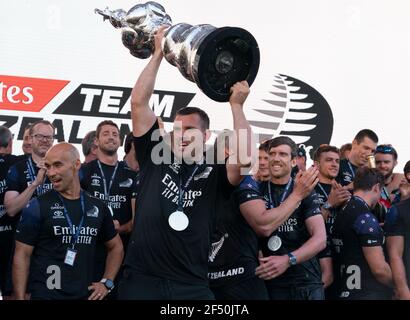 Auckland, Neuseeland, 17. März 2021 - Emirates Team der neuseeländische Grinder Joe Sullivan hält während der Siegerehrung den America's Cup nach der Niederlage der italienischen Herausforderer Luna Rossa Prada Pirelli. Hinten rechts ist Skipper Peter Burling mit anderen Crew-Mitgliedern feiern. Kredit: Rob Taggart/Alamy Stockfoto