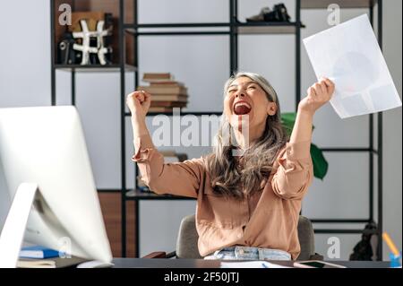 Glückliche erfolgreiche einflussreiche reife asiatische Frau, Geschäftsfrau, ceo, sitzt in einem modernen Büro, trägt stilvolle Kleidung, freuen sich über Erfolg, Gewinn, Sieg, Mit Händen gestikulieren, lächeln Stockfoto