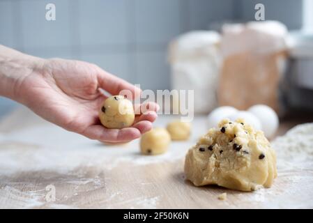 Weibliche Hand hält eine Kugel Plätzchenteig, ein Stück Teig mit Schokolade auf dem Küchentisch, Kochen Cookies zu Hause, Nahaufnahme. Stockfoto