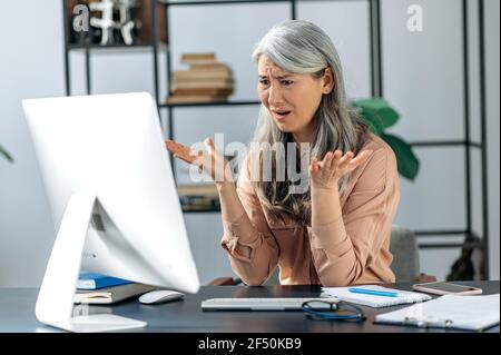 Empört verärgert schöne asiatische reife Frau, Geschäftsfrau oder Manager, schaut auf Computer in Verwirrung, schreit, erhält unerwartete Nachrichten, scheitert den Deal, Gesten, Sitzt am Tisch im Büro Stockfoto