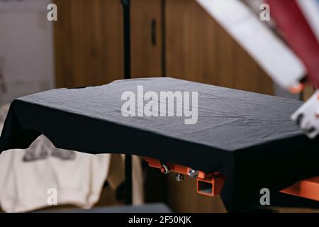 Selektiver Fokus Foto von blanken schwarzen T-Shirt auf Holzregal auf dem Druck Siebgerät. Siebdruck Produktion. Drucken von Bildern auf T-Shirts von Stockfoto