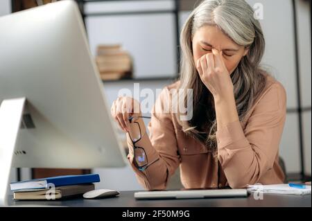 Überfordert müde traurige ältere grauhaarige Asiatin, Freiberuflerin, Managerin, hält Brille in der Hand, in stilvoller Kleidung, Kopfschmerzen, Stress nach langer Computerarbeit, massieren der Augen am Arbeitsplatz Stockfoto