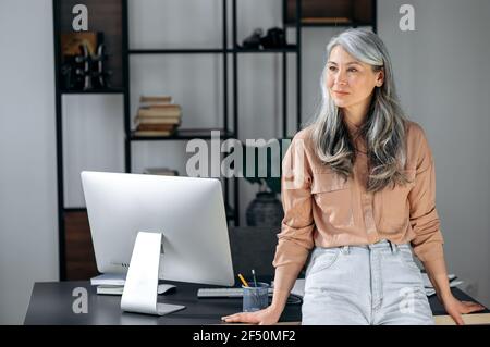 Porträt einer selbstbewussten erfolgreichen reifen grauhaarigen asiatischen Frau, Geschäftsfrau, Führungskraft oder Coach, die in einem modernen Büro in der Nähe des Arbeitstisches steht, in stilvoller Kleidung trägt, zur Seite schaut und lächelt Stockfoto