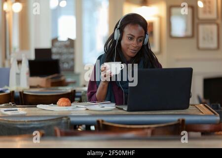 Junge Frau mit Kopfhörern, die Kaffee am Laptop im Café trinkt Stockfoto