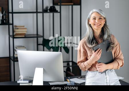 Porträt einer selbstbewussten, schönen, smarten, grauhaarigen asiatischen Geschäftsfrau oder Geschäftsführerin, die im modernen Büro steht, in stilvoller Kleidung gekleidet ist, Ordner in den Händen hält, die Kamera anschaut und freundlich lächelt Stockfoto