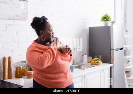 Zufrieden african american plus size Frau hält Kunststoff-Tasse und Trinken leckeren Smoothie in der Küche Stockfoto
