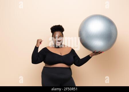 Fröhliche afroamerikanische plus size Frau in Sportbekleidung halten Fitness Ball isoliert auf beige Stockfoto