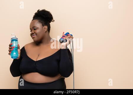 Happy african american plus size Frau in Sportswear Holding Sport Flasche und Springseil isoliert auf beige Stockfoto