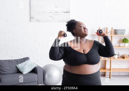 Lächelnd african american plus size Frau in Sportswear Ausübung mit Kurzhanteln im Wohnzimmer Stockfoto
