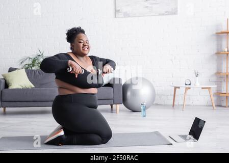 Lächelnd afrikanisch plus size Frau Ausübung mit Hanteln in der Nähe Laptop in der Nähe der Fitnessmatte Stockfoto