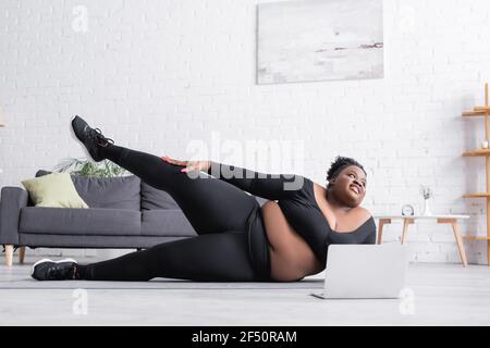 Fröhliche afrikanisch-amerikanische plus size Frau Ausübung in der Nähe Laptop auf Fitnessmatte Stockfoto
