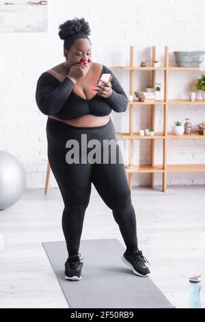 Überrascht afroamerikanische plus size Frau in Sportkleidung Blick auf Smartphone und Mundbereich Stockfoto