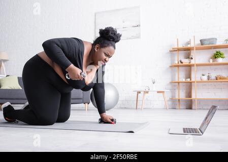 Angespannte afroamerikanische plus size Frau in Sportswear Ausübung mit Kurzhanteln und Blick auf Laptop Stockfoto
